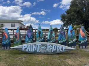 Paint and Paddle - Mon Forest Towns - Mon Forest Towns Row of tall, colorful canoe kiosks on a fence, each featuring illustrations of people; a painted boat lies on the ground in front, with a white house and trees in the background.